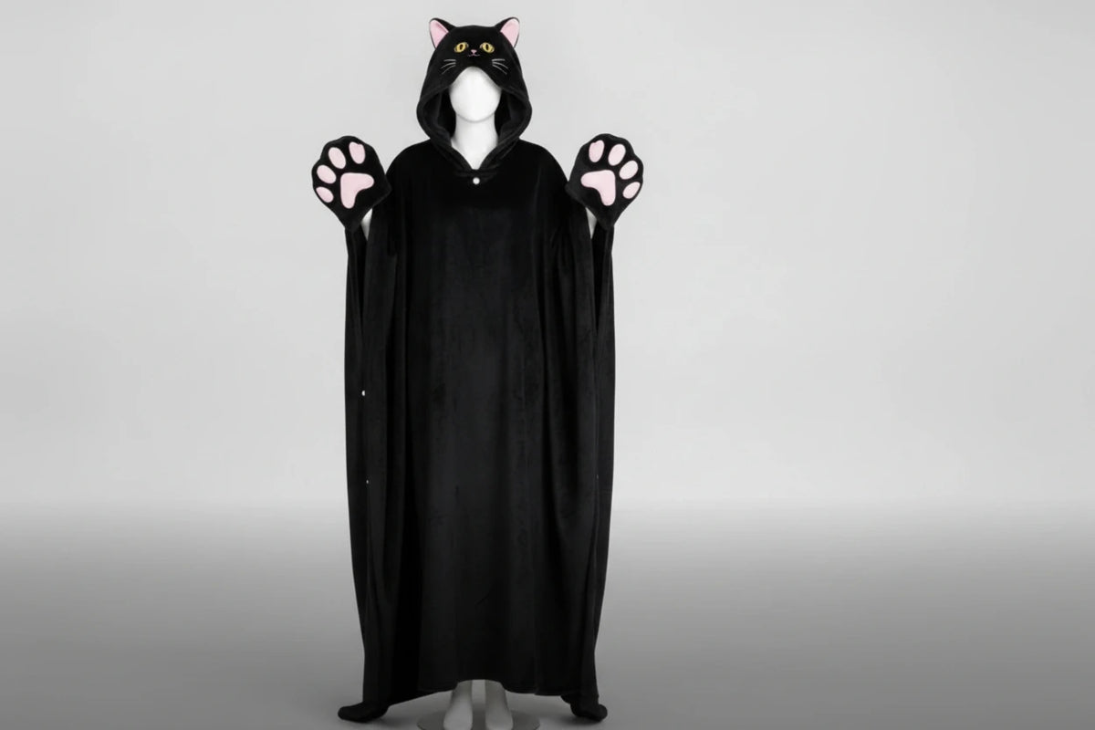 A black cat-themed hooded blanket/cape wear garment displayed on a white mannequin against a light gray studio background. The hood has pink inner ears and yellow eyes, and the glove-like hands have pink paw pads.