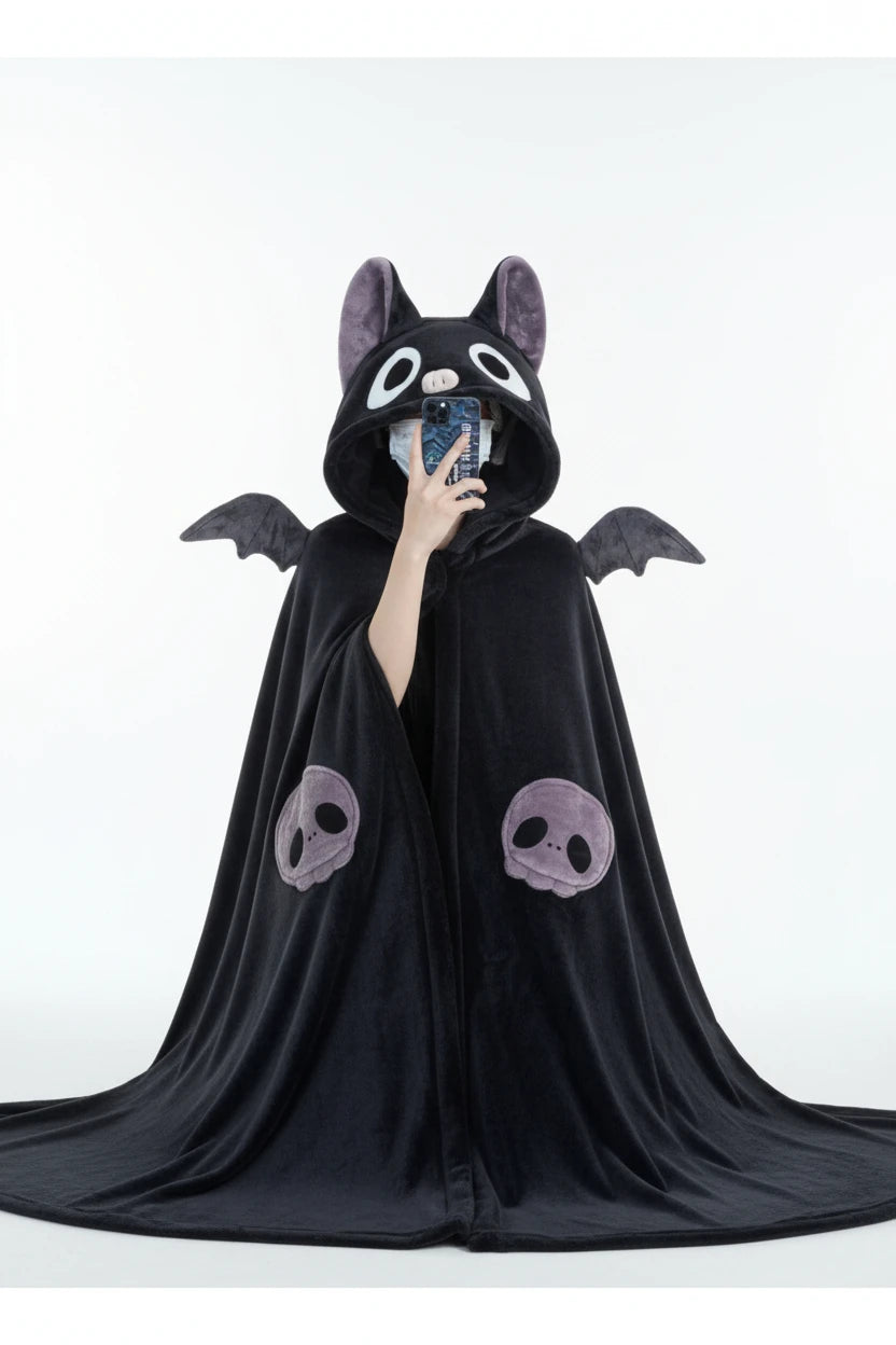 A person wearing a plush black bat-themed hooded blanket or cape with gray wings, large white eyes, and purple skull details on the front, taking a mirror selfie in a room.
