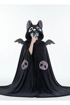 A person wearing a plush black bat-themed hooded blanket or cape with gray wings, large white eyes, and purple skull details on the front, taking a mirror selfie in a room.