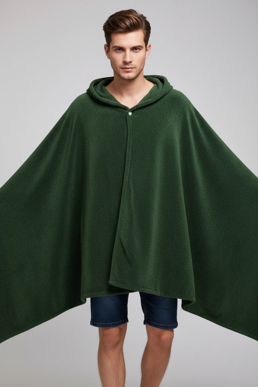 A woman wearing a dark green hooded blanket cape with a drawstring closure, standing in a brightly lit room with a light blue wall and a lamp in the background.
