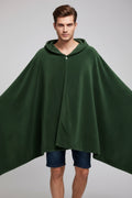 A woman wearing a dark green hooded blanket cape with a drawstring closure, standing in a brightly lit room with a light blue wall and a lamp in the background.