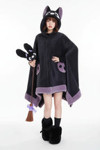 A person wearing a plush bat-shaped hooded blanket or cape in dark charcoal gray with lavender trim, seated on a white cushion in a stylized room with purple walls and a black bat-shaped decor piece.