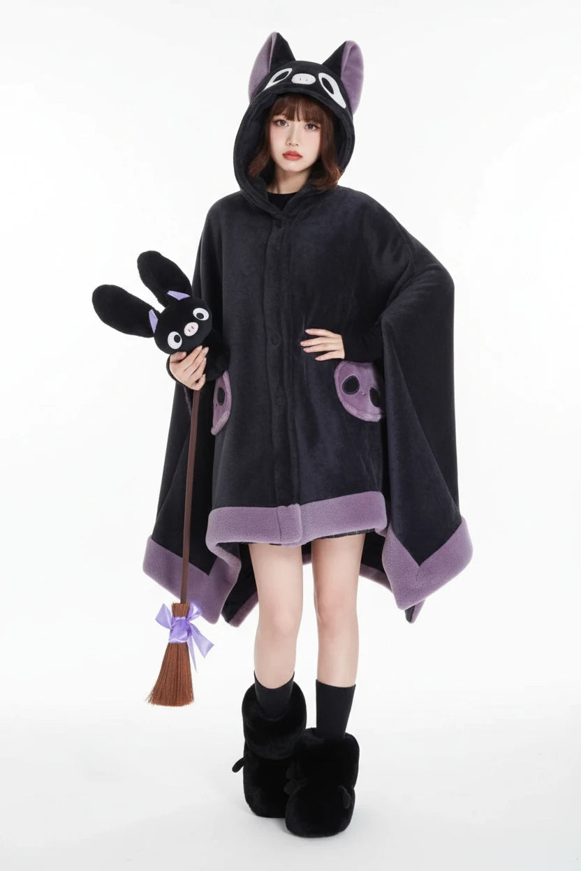 A person wearing a plush bat-shaped hooded blanket or cape in dark charcoal gray with lavender trim, seated on a white cushion in a stylized room with purple walls and a black bat-shaped decor piece.