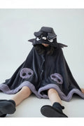 A person sitting on a wooden floor wearing a plush dark gray hooded blanket or robe with cartoon face details and matching cuffs, in a cozy bedroom setting.