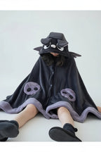 A person sitting on a wooden floor wearing a plush dark gray hooded blanket or robe with cartoon face details and matching cuffs, in a cozy bedroom setting.