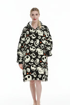 A person wearing a hooded blanket poncho with an all-over Halloween-themed print featuring Jack Skellington skulls, ghosts, bats, and small orange diamond accents on a black background, against a plain white studio backdrop.