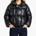 A model wearing a shiny black cropped puffer jacket with a high collar and horizontal quilting, paired with dark blue jeans, against a plain white background.